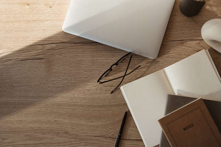 Home office desk workspace with laptop, notebook, glasses, sunlight shadows on neutral wooden...