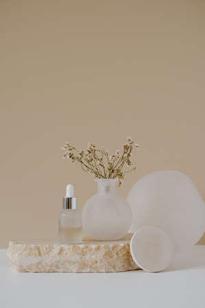Aesthetic minimalist beauty care therapy concept. Organic serum oil cosmetics bottle on stone with flowers against neutral beige background. Body skin, face treatment product compositionの写真素材