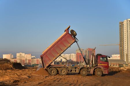 Red dump truck dumps its load of sand and soil on construction site for road construction or for foundation work. Transportation of bulk cargo.  Trucking industry, freight cargo transportの写真素材