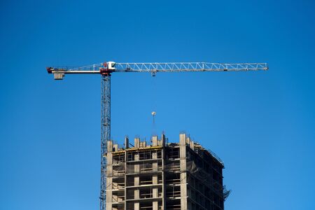 Tower crane lifts the block for building under construction at background blue sky at sunny day. Precast concrete  slab hanging from crane hook above building skeleton at construction siteの写真素材