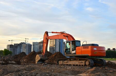 Crawler excavator in construction site on the background of tower cranes and multi-storey residential buildings and shopping centers under construction. Special heavy equipment on blue sky backgroundの写真素材