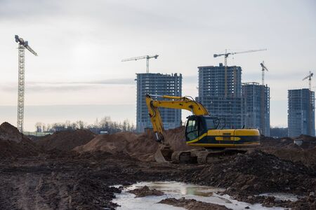 Yellow excavator with bucket at building under construction. Backhoe at large scale construction site against tower cranes. Digging the ground for the foundation and for laying sewer pipesの写真素材