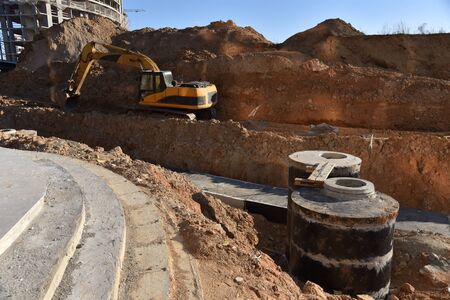 Excavator on earthwork during the laying of pipes of the heating system to a new residential building at the construction site. Laying concrete sewer wells, rings and manholes in ground.の写真素材