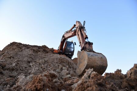 Excavator during earthmoving work at open-pit mining on sunset background. Loader machine with bucket in sand quarry. Backhoe digging th ground for the foundation and for laying sewer pipesの写真素材