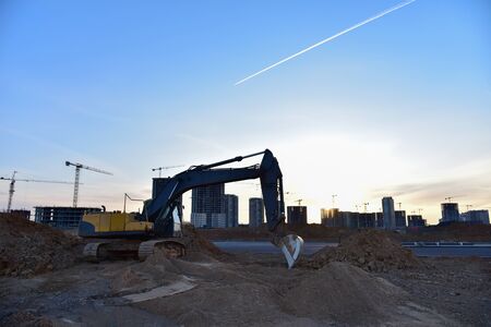 Excavator and tower crane at construction site on sunset background. Cranes constructing a new...