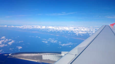 Airplane passenger window view of wing in flight with clouds on blue sky background. Small roughness...