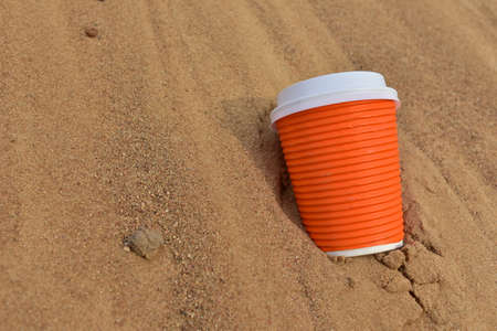 Discarded Paper coffee cup on sand at beach. Disposable coffee cup on sandy background. The problem of environmental pollution. Pile of abandoned garbage, including food waste, fast food packagingの写真素材