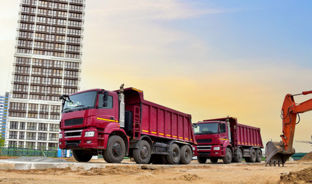 Dump truck and excavator working on earthworks at construction site on sunset background. Trucks for transportation of bulk cargo. Trucking industry, freight cargo transport conceptの写真素材
