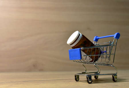 Pills in bottle in shopping cart on wooden background. Medicine grade pharmaceutical tablets at mini shopping trolley. Medical pill for maintaining and improving health in shopping basket cartの写真素材