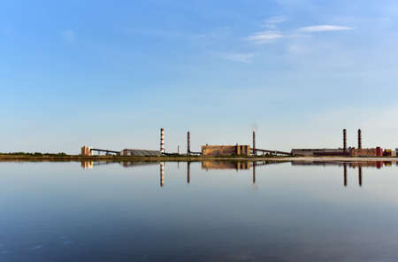 View of an industrial plant with chimneys and workshop buildings near the lake against blue sky. Cleaning Up after pollution. Safe drinking water foundation. Release of toxic substances into the airの写真素材