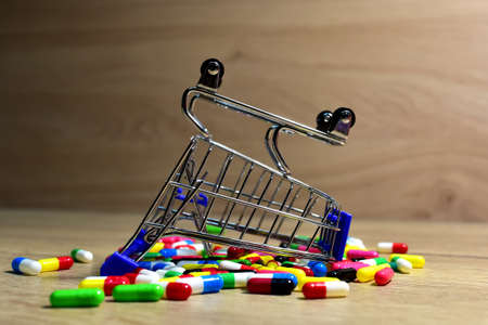 Multicolored pills in capsules and inverted shopping cart on wooden a table. Medicine grade pharmaceutical tablets and mini shopping trolley. Medical pill for maintaining and improving health.の写真素材