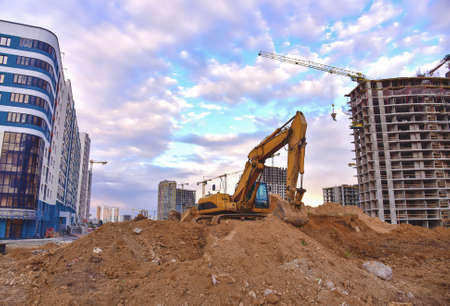Excavator on earthworks at construction site. Backhoe on road work and laying sewer pipes. Construction machinery for dig ground, excavating foundation. Tower cranes on building construction.の写真素材