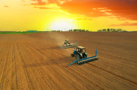 Tractor sowing seed on plowed field. Sowing seeds of corn and sunflower. Blue Tractor with disk harrow on plowing field. Seeding machinery on farm field. Seed sowing in farmland on sunset.の写真素材