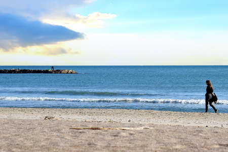 Girl is walking near the sea. Young woman walks along the sand of a beach near Mediterranean Sea against backdrop of waves and a stormy sky at sunset. Female person walks along coastline of the ocean.の写真素材