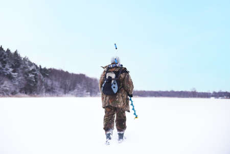 Ice fishing on frozen lake. Fishermen in camouflage clothing is walking on frozen lake for ice fishing. Fisherman on lake with drill for drilling holes in the ice for catching fish. winter fishing.の写真素材