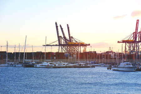 Container crane in seaport on sunset. Yachts and motor boats in La Marina de Valencia. Unloading sea martime containers. Container ship and shipping containers box. Luxury yacht and Sailboat in port.の写真素材