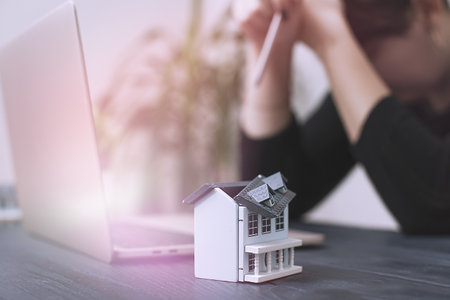 Businesswoman Facing Challenges in Real Estate Investment: Model House on a Desk with Laptop in a Modern Workspace Settingの写真素材