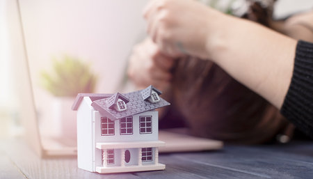 Businesswoman Facing Challenges in Real Estate Investment: Model House on a Desk with Laptop in a Modern Workspace Settingの写真素材