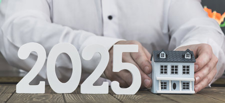 2025 Real Estate Vision: Hands Holding a Model House with Stock Market Charts in the Background for Futureの写真素材