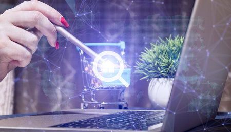 Digital Real Estate Search and E-Commerce Workspace with Hand Using Stylus on Shopping Cart Icon, Laptop with Network Graphics, Green Plant, and Modern Office Desk Settingの写真素材