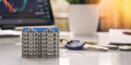 Miniature building model with house keys on a desk, financial stock chart in background, symbolizing property investment and housing market.の写真素材