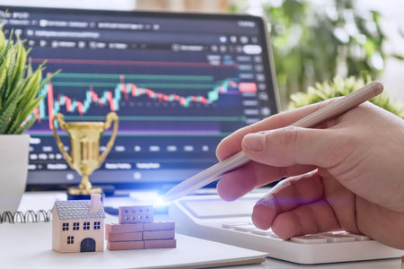 Hand holding stylus points to miniature buildings with stock chart in background, representing real estate investment and digital analysis.の写真素材