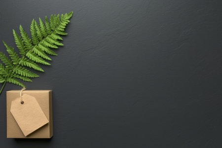 Small eco-friendly gift box wrapped in kraft paper with blank tag, decorated with a green fern leaf on a dark surface.の素材