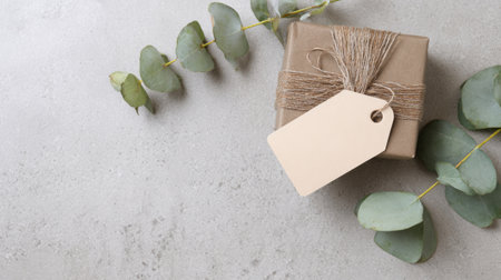 Elegant wrapped gift box decorated with rustic twine, blank kraft paper tags, and fresh greenery on a textured concrete background.の素材