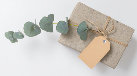 Elegant wrapped gift box decorated with rustic twine, blank kraft paper tags, and fresh greenery on a textured concrete background.の素材