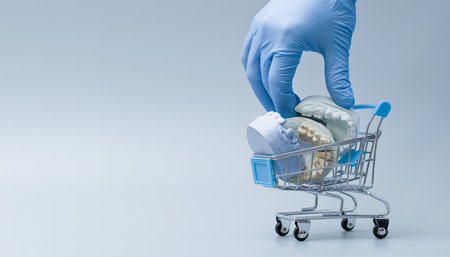A gloved hand places dental jaw models into a miniature shopping cart, symbolizing oral care, dental services, and healthcare decisions.の写真素材