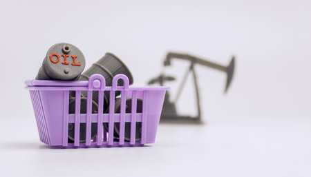 A small shopping basket filled with miniature oil barrels with a blurred pumpjack in the background,...