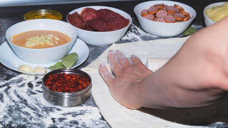 Pizza ingredients. hand rolls out the dough. rolling pin and flour. sliced salami and sausages. grated cheese and pizza sauce with spicesの写真素材