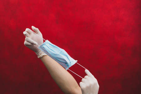Hands in protective gloves stretch a medical mask on a red background. preventive measures against respiratory illness and virus. hands in white surgical gloves holds disposable mask for protectionの写真素材
