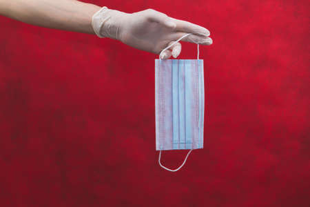 Blue medical mask hanging on fingers. hand in white surgical glove holds protective mask. red background with copy spaceの写真素材