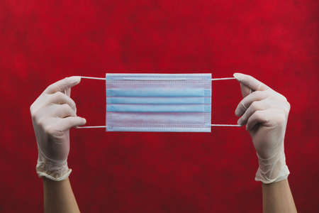 Blue medical mask in hands with white gloves on a red background. preventive measures against respiratory illness and virus. hands in white surgical gloves holds disposable mask for protectionの写真素材
