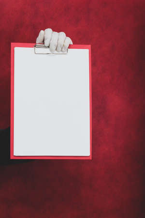 Hand in surgical glove holding white blank paper sheet on red background. template mock up. surface of leaflet. copy spaceの写真素材