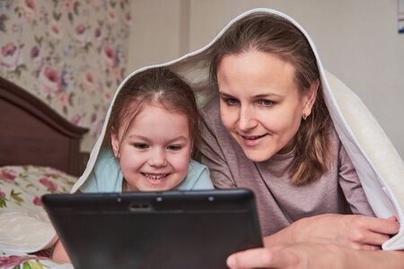 A charming little girl and her beautiful young mother use a digital tablet and smile while sitting on chairs at homeの写真素材