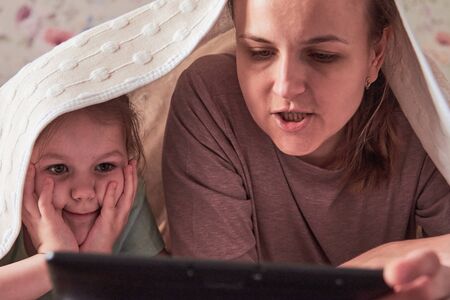 A charming little girl and her beautiful young mother use a digital tablet and smile while sitting on chairs at homeの写真素材