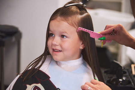 a little girl in a barbershop. Childrens haircut, hair drying, combing and styling. The child is happy and contented. Barber station wagon.の写真素材