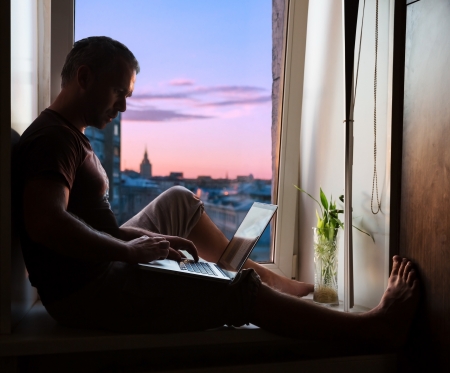 Silhouette of man sitting on window sill with laptop on background of cityscape at sunsetの写真素材