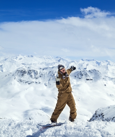 Cheerful snowboarder in protective mask showing sign on the background beautiful mountains and blue sky with cloudsの写真素材