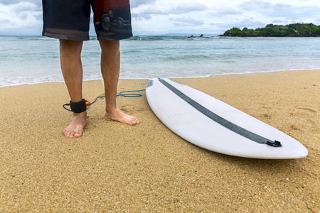 Lower half of surfer on sand, standing on the beach with an attached surfboard near the oceanの写真素材