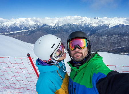 Happy couple snowboarders standing on edge of mountain peaks and taking selfie portrait with camera or smartphone on  background of snowy mountains in ski resortの写真素材