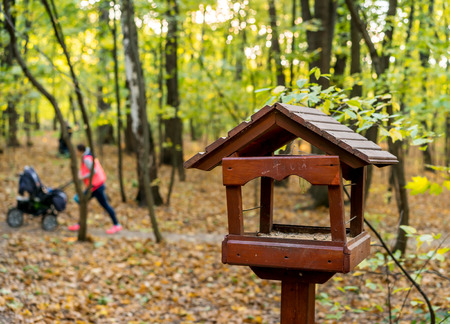 Wooden bird feeder on pole stands in autumn forest park. In background, mother walking with baby in strollerの写真素材