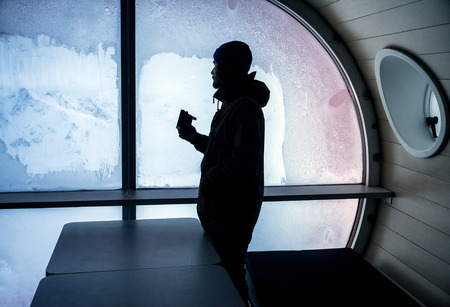 Bearded man traveler inside the hotel with flask in his hand looking out the window at the beautiful snow-covered mountain landscapeの写真素材