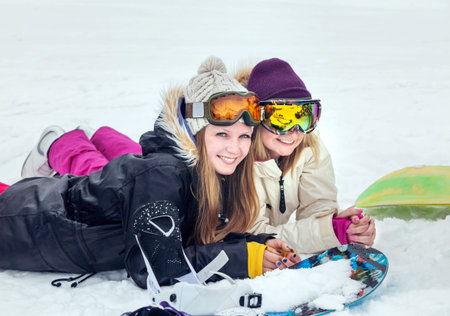 Two positive young women snowboarders lie on snow in slope mountains on winter resort