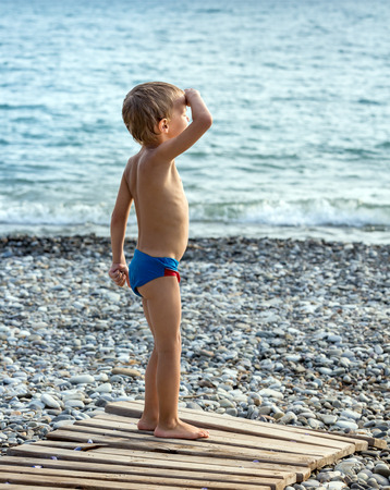 Young boy stands on the beach, raising his hand to his forehead looking into the distanceの写真素材