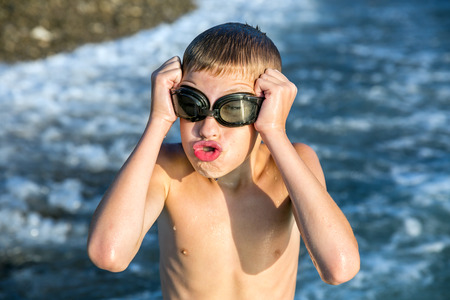 Young funny boy with braces on his teeth, shows tongue and grimace on his face with glasses for swimming on sea beachの写真素材