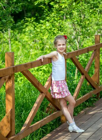 Portrait of beautiful young positive girl in park with braids and red flower in summertime on background of green vegetation in the park near of wooden fenceの写真素材