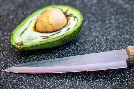 Half an avocado fruit, cut up, lies on the table with kitchen knife closeup on the background